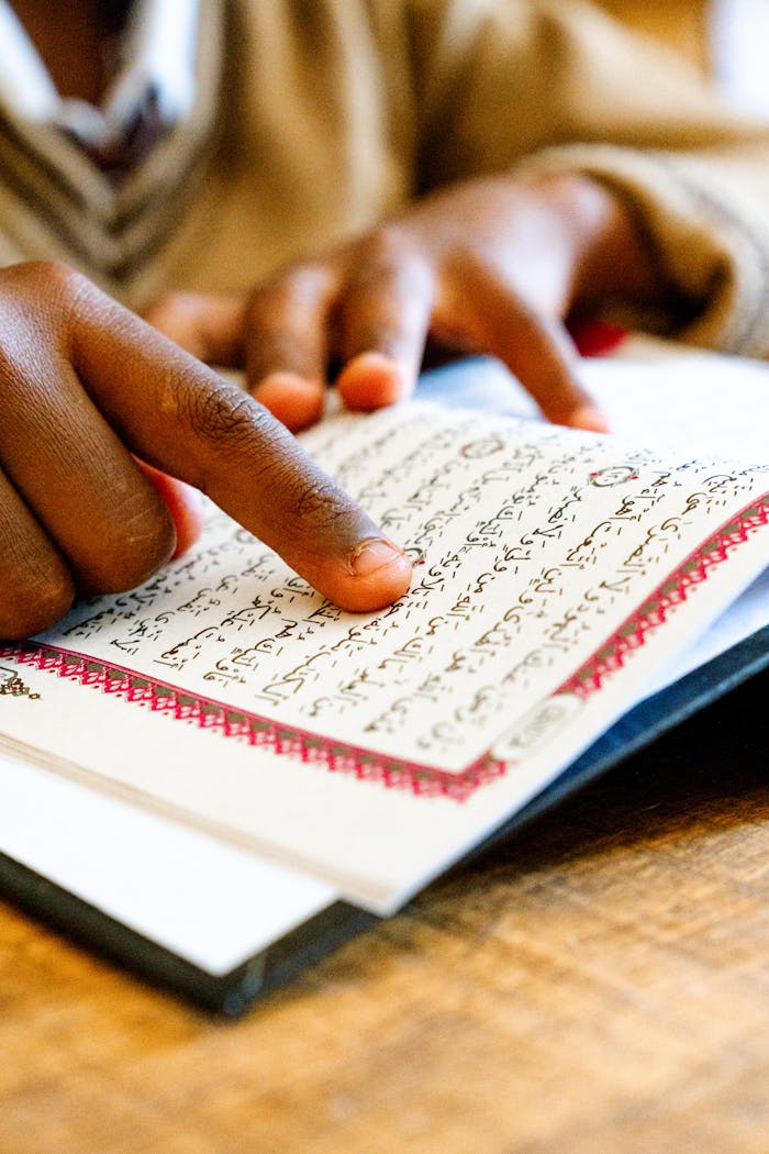 Close-up of a child's hand pointing while reading the Quran indoors.