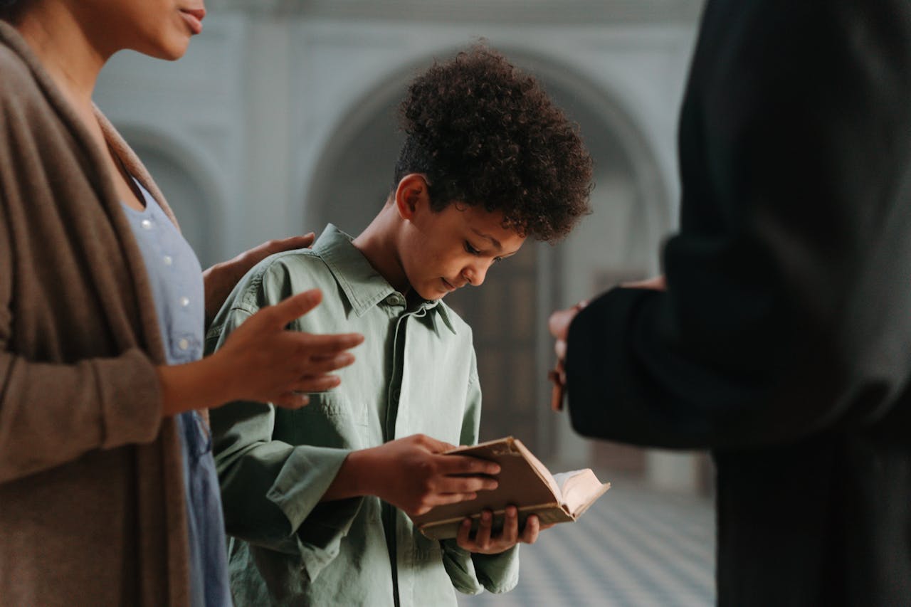 services-04 A young boy reads a religious book with guidance from adults in an indoor church setting.